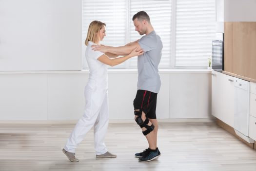 Young Man Trying To Walk With A Female Physiotherapist In Clinic