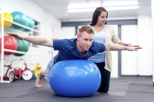 Adult male patient balancing on a therapy exercise ball during a therapy session while an adult female physical therapist helps maintain his balance