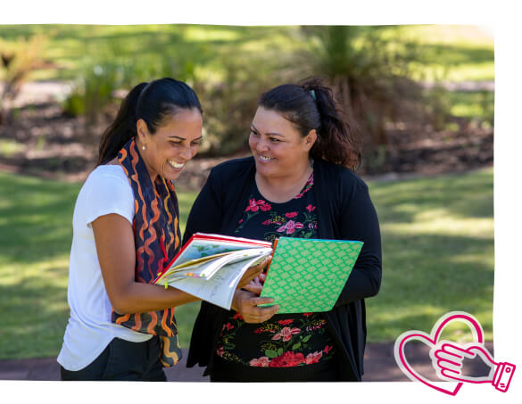 Two women smiling and talking in a park while they look at a book